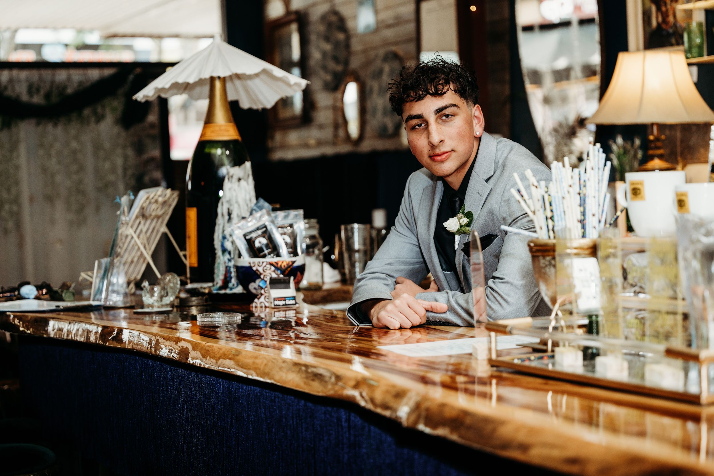 A person in a blue button-down shirt leans casually on a richly-textured wooden bar counter surrounded by glassware.