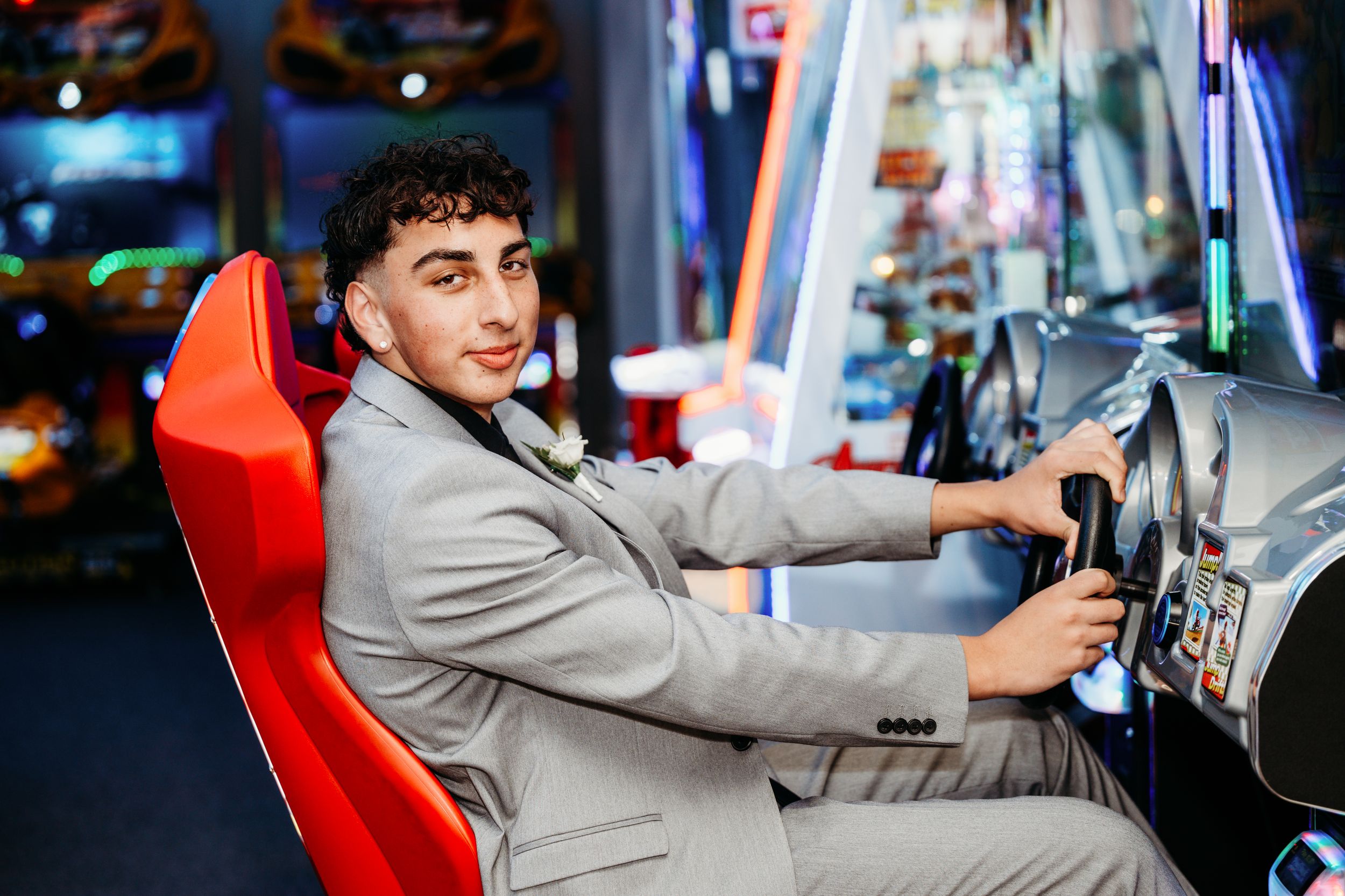A person in a grey suit enjoys playing an arcade racing game while seated in a red gaming chair.