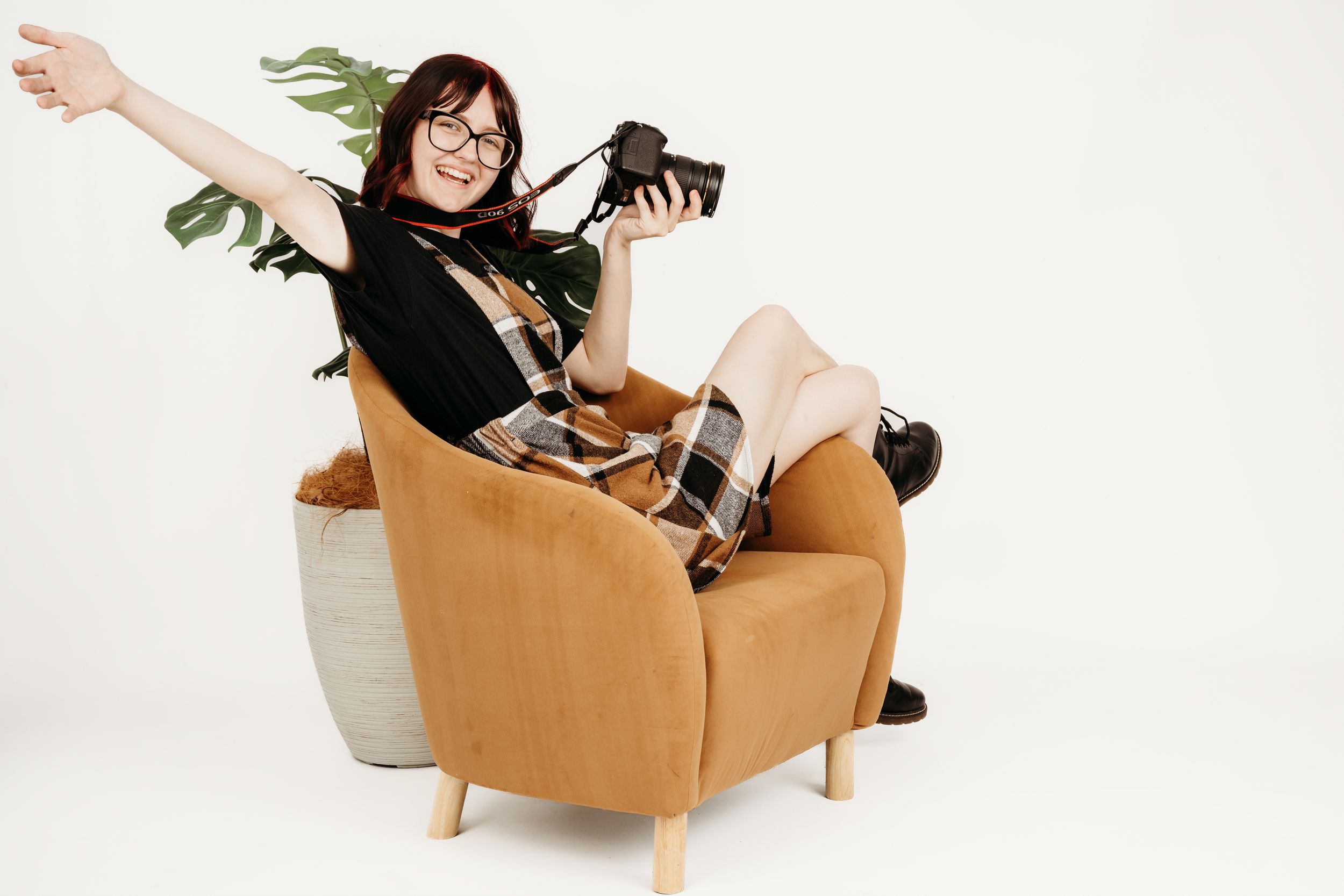 Smiling woman with red hair and glasses sitting in orange armchair holding camera with arm outstretched, white background.