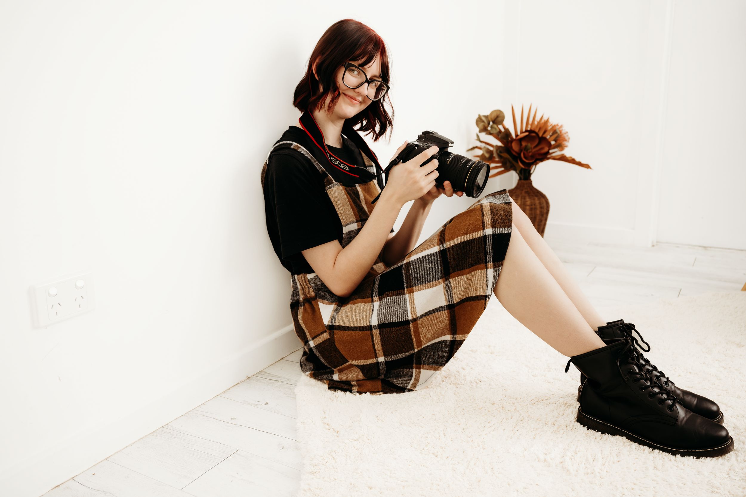 Young woman with red hair and glasses sits on floor holding a camera and dried flowers, wearing plaid dress and black boots.