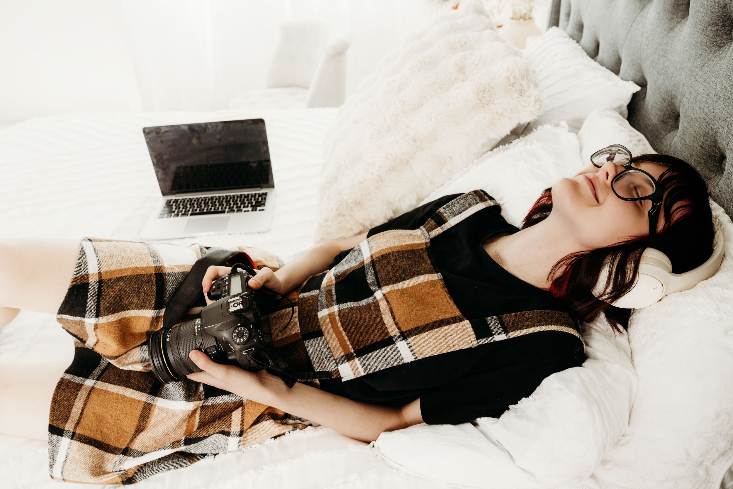 Young woman with glasses lying on bed holding a DSLR camera beside an open laptop, wrapped in a plaid blanket.