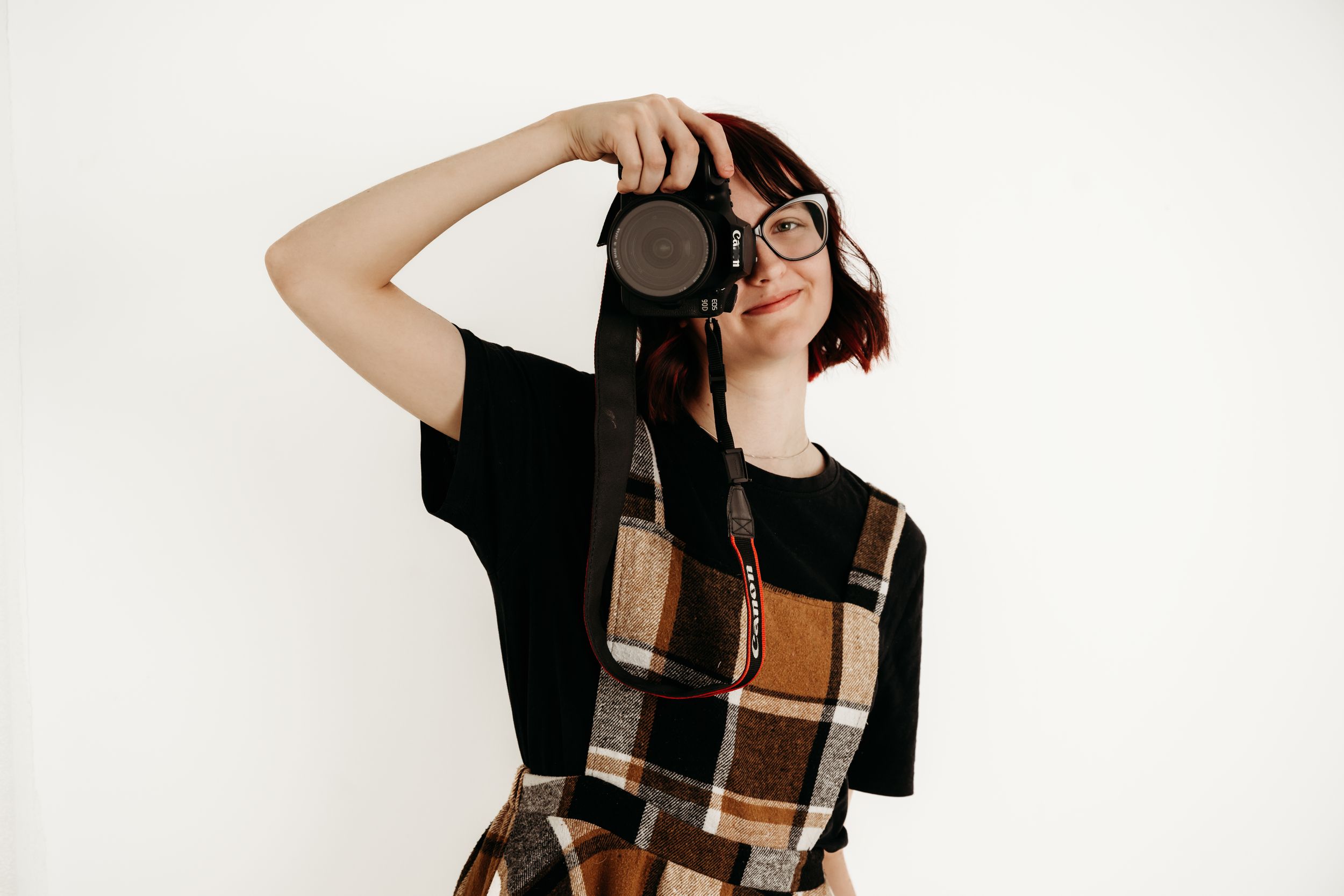 Young woman with glasses and plaid overalls holds a DSLR camera up to her face against a white background.
