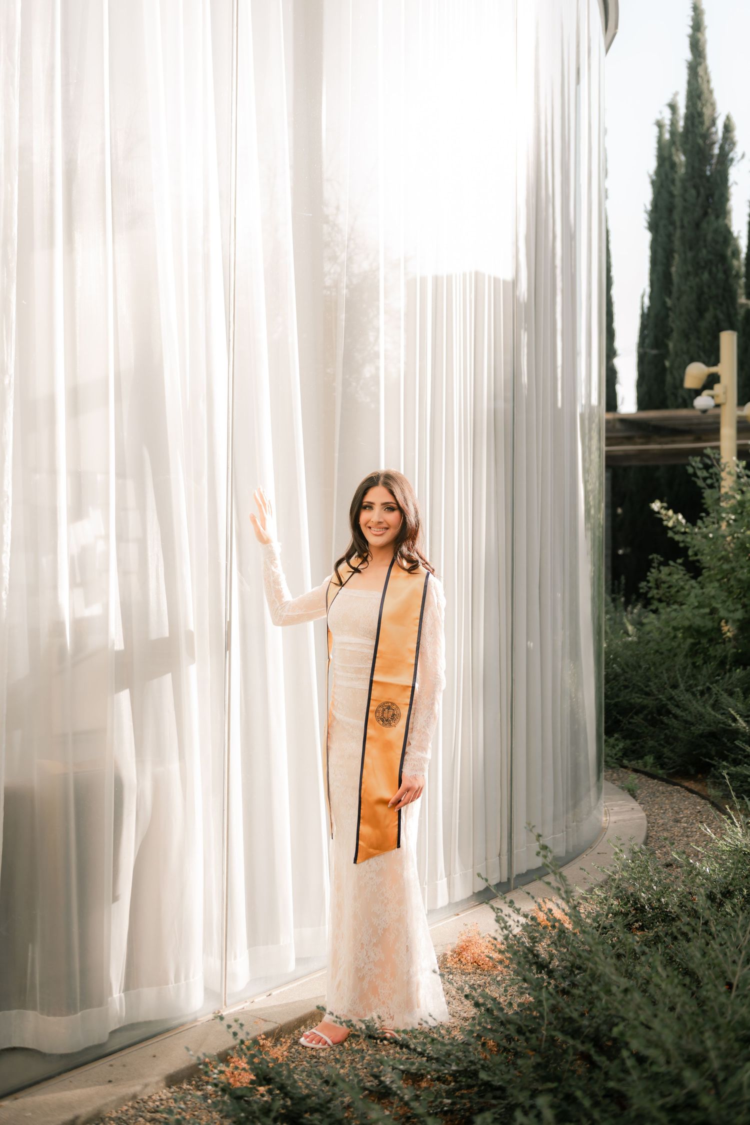 A UC Davis graduate poses next to a big glass wall on the UC Davis campus. Photographed by Shumesa Mohsin