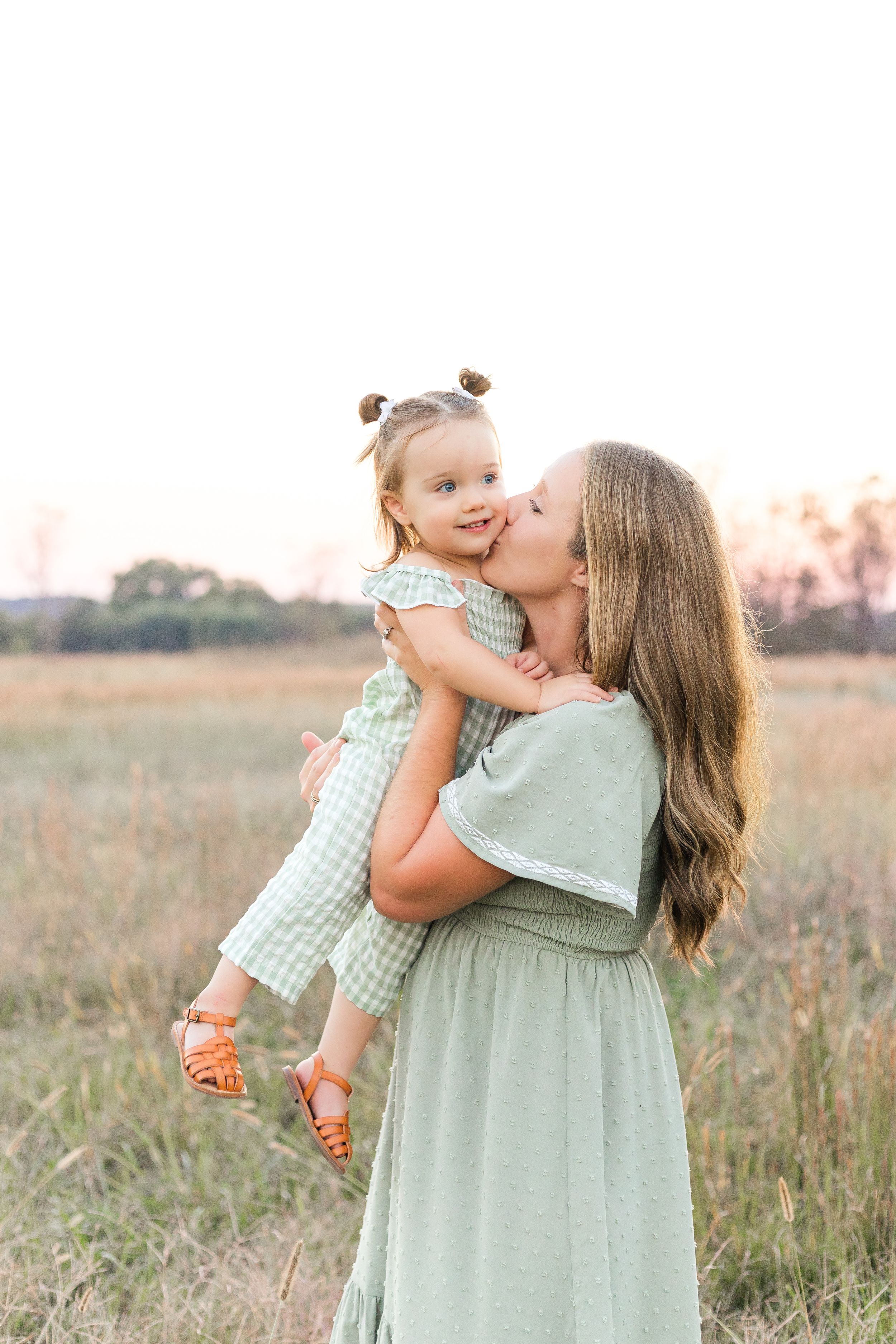 Mother wearing a sage green dress holding her young child close in a grassy field during a warm outdoor family portrait session.