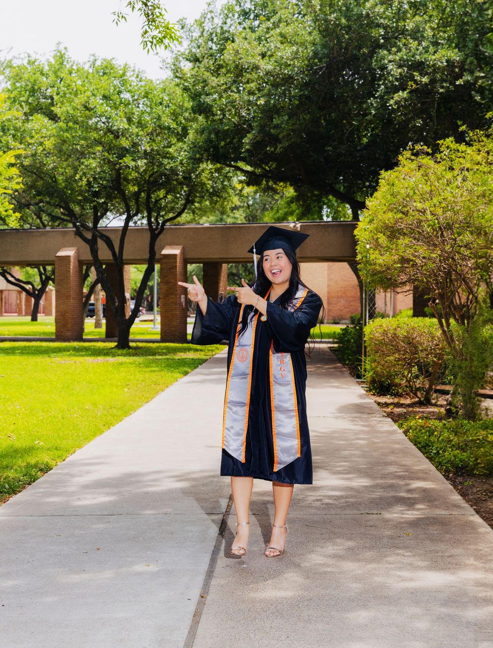 Graduate in navy blue gown poses on campus walkway surrounded by greenery.