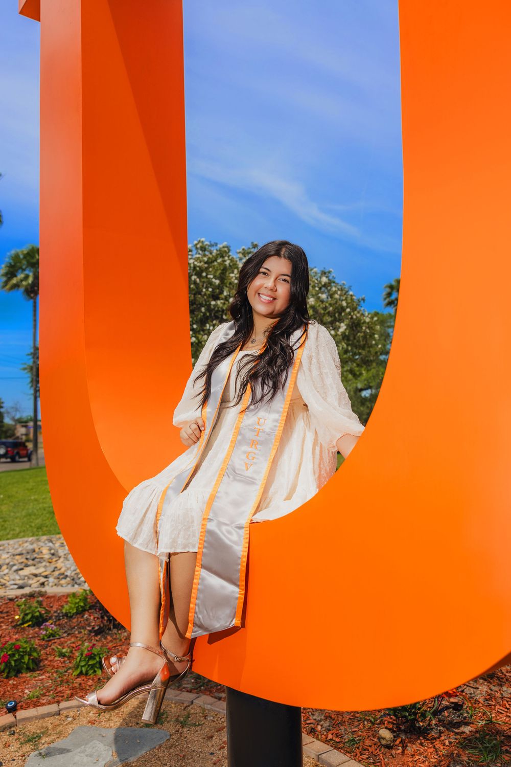 Woman in white dress poses against vibrant orange U-shaped sculpture outdoors.