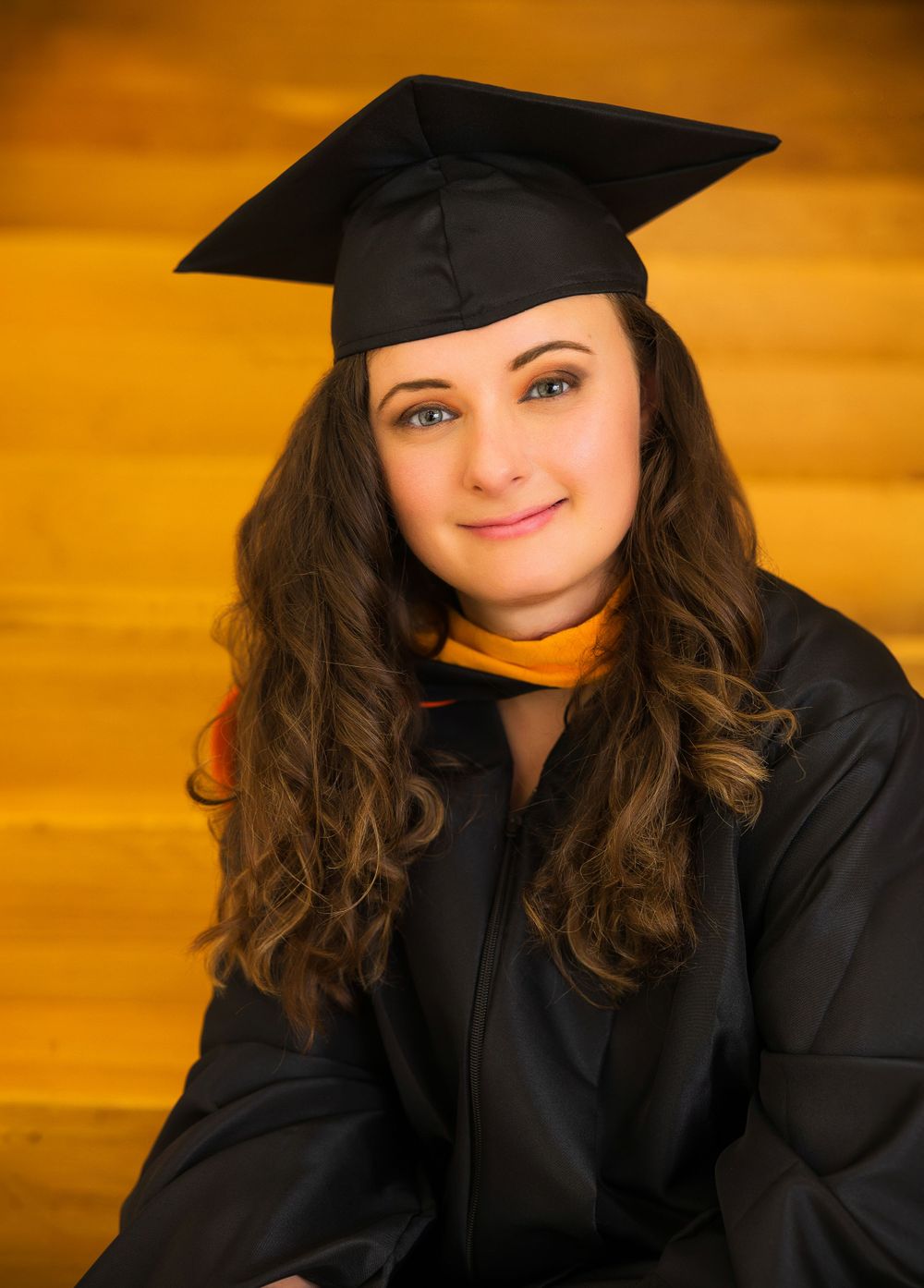 Graduate in black cap and gown poses against warm yellow background.