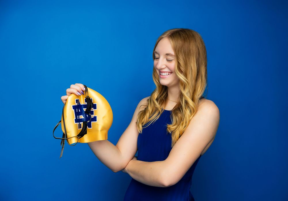 Athletic swimmer in navy suit holds yellow swim cap against vibrant blue background.