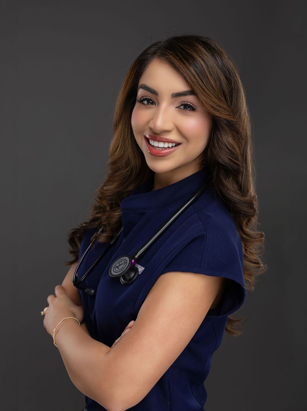 Female healthcare professional in navy scrubs with stethoscope smiling confidently with arms crossed.