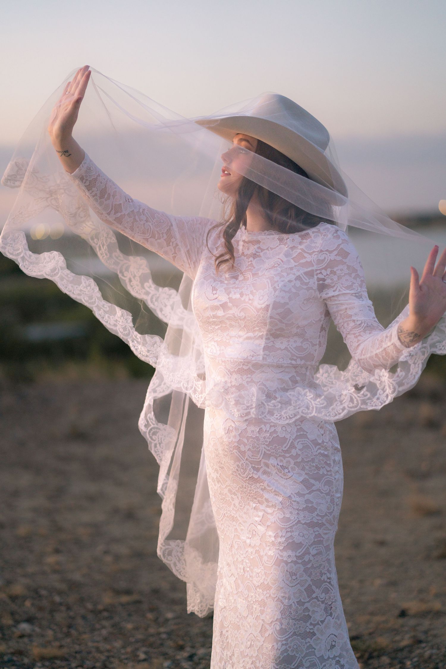 A figure in a white lace dress and hat twirls gracefully on a beach at sunset.