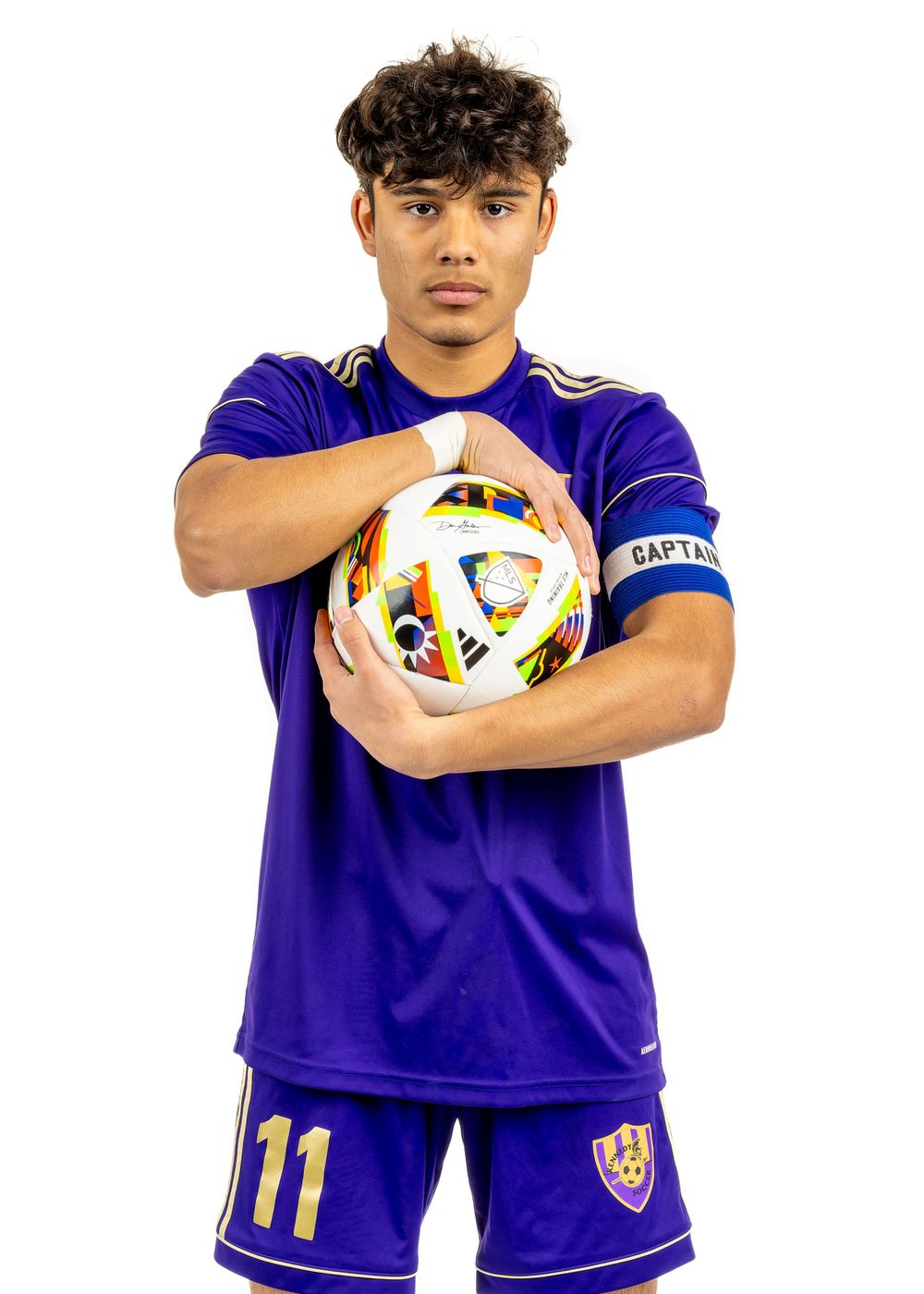 Soccer player in purple uniform holds ball against plain studio backdrop.