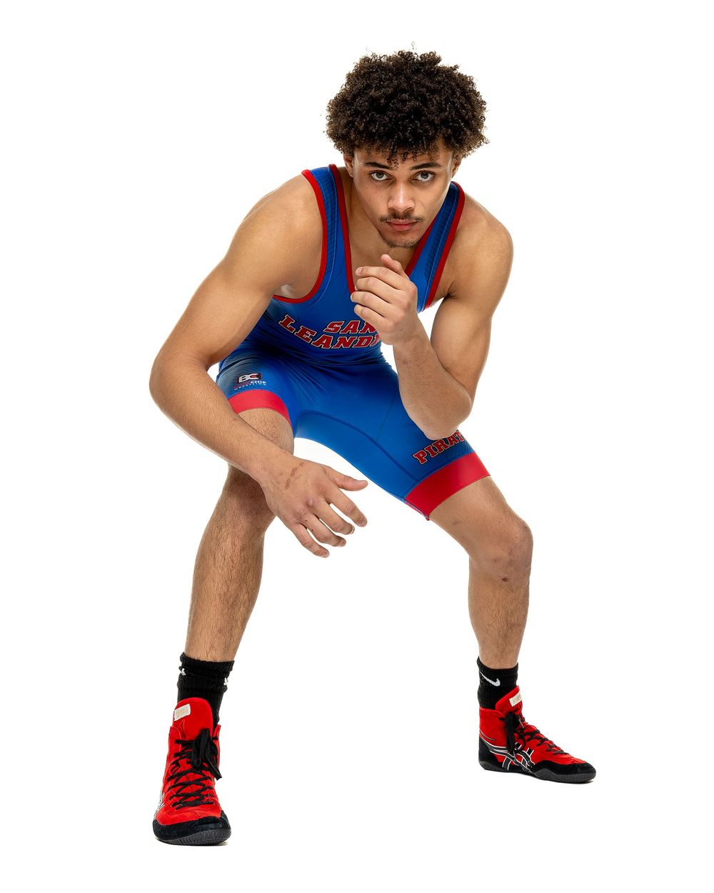 Wrestler in blue and red singlet poses in ready stance against white background.
