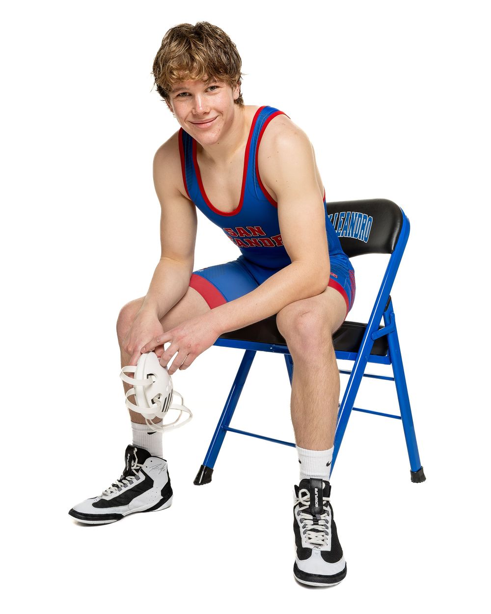Wrestler in blue and red singlet sits on folding chair against white background.