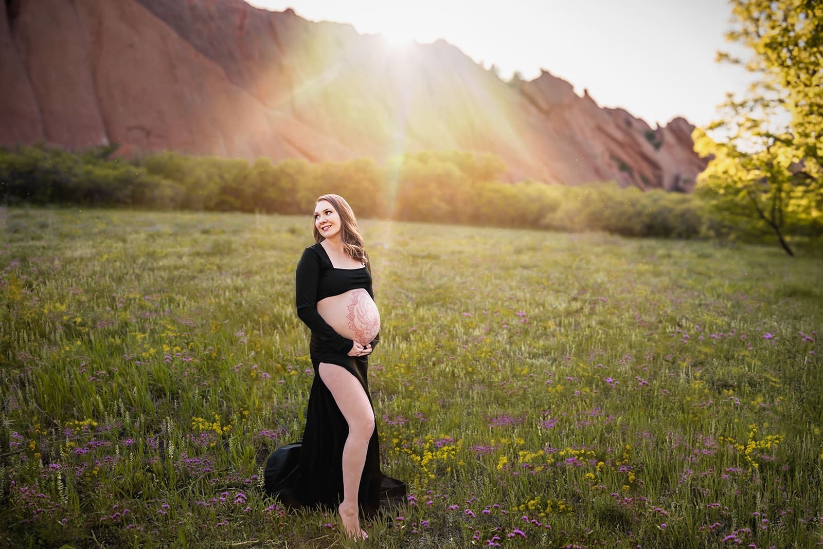 Sunlit maternity portrait in black gown at Red Rocks meadow, surrounded by wildflowers, by Denver maternity photographer.