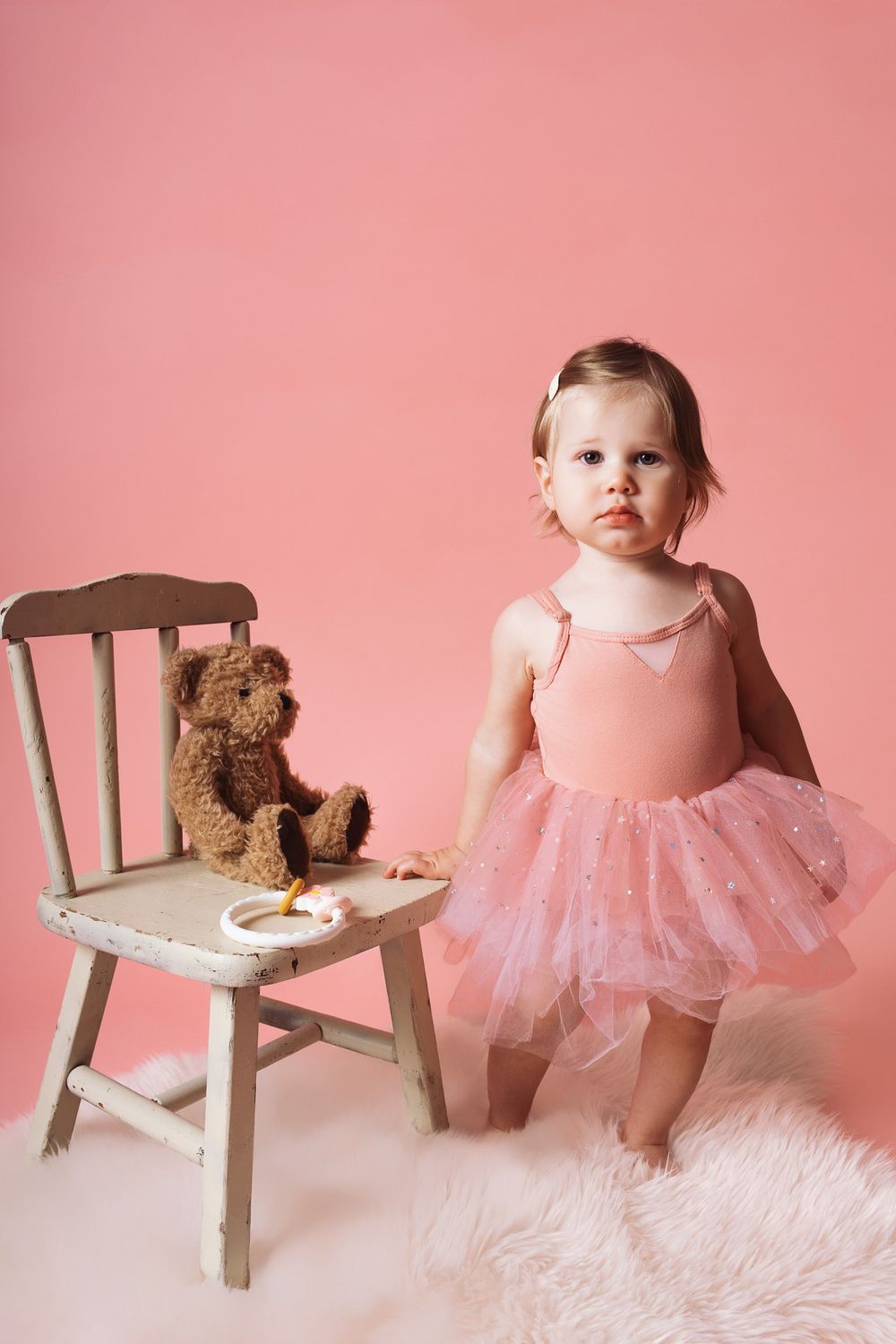 A series of milestone photos showing a baby in a pink tutu with a teddy bear and wooden chair against a pink backdrop.