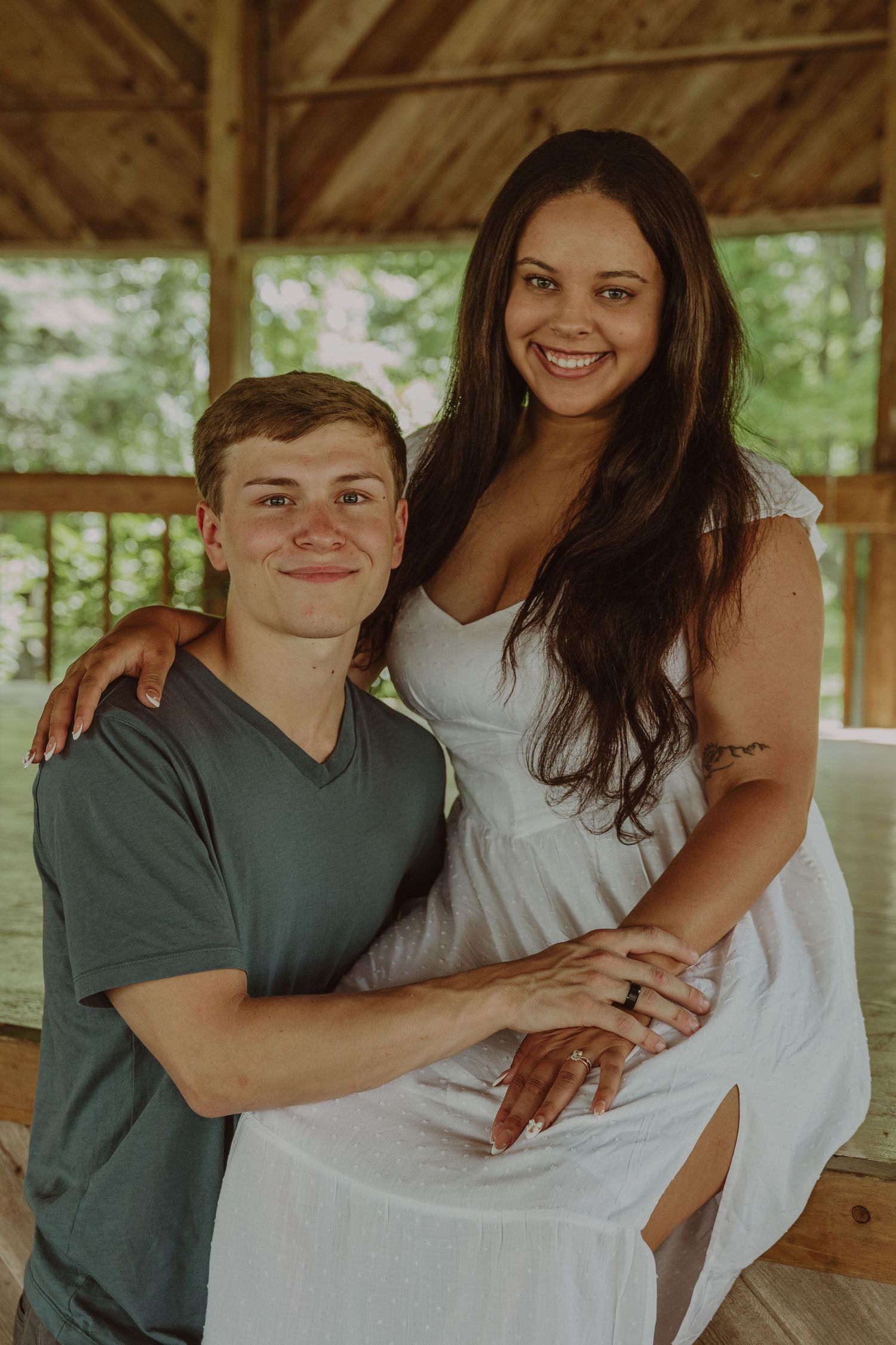 A couple poses together in a wooden gazebo, with the woman in a white dress and the man in a dark shirt.