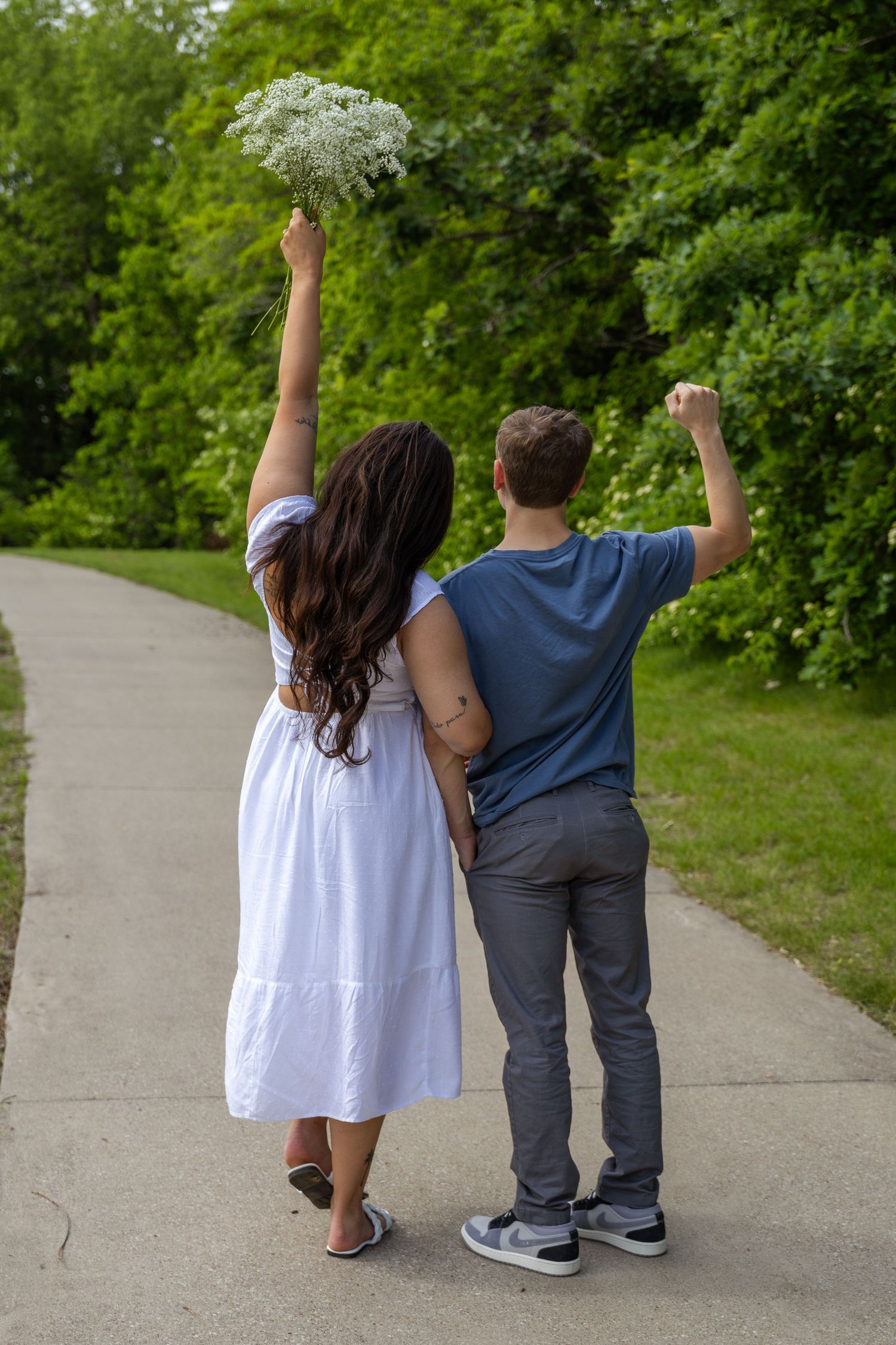 A couple celebrates with raised arms while walking on a path, holding white flowers.