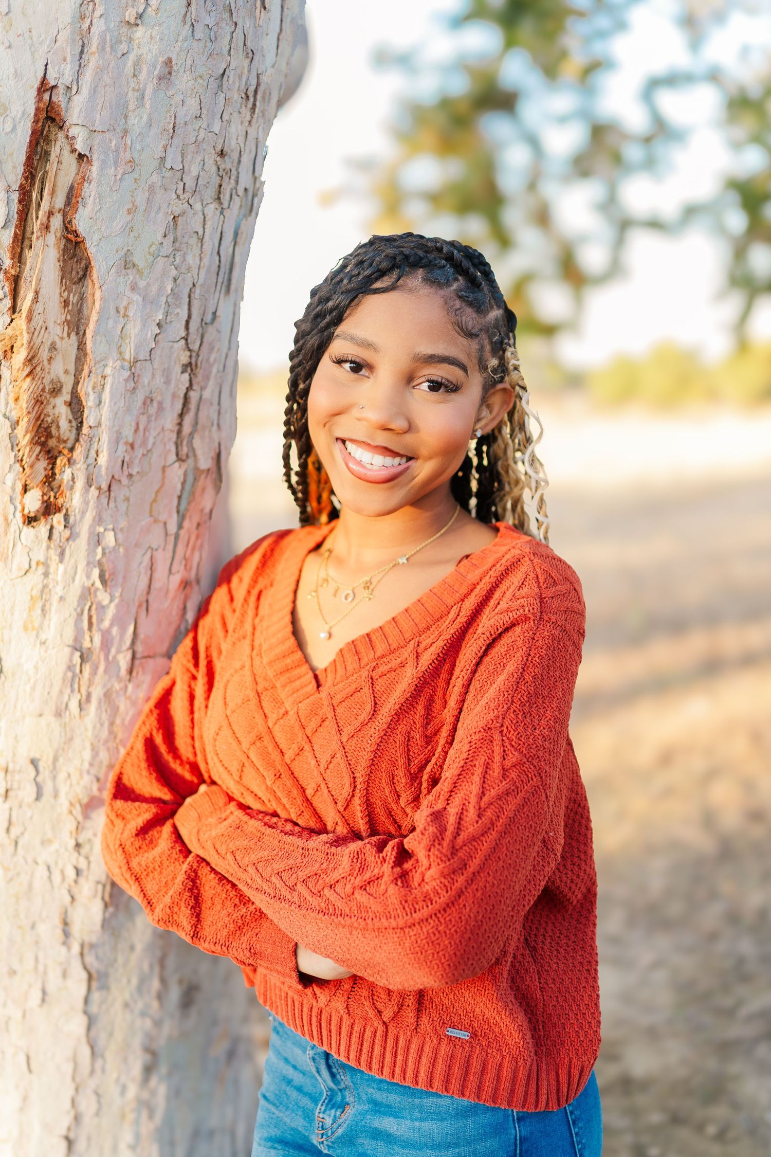 A person in an orange sweater poses against a tree trunk in warm sunlight.