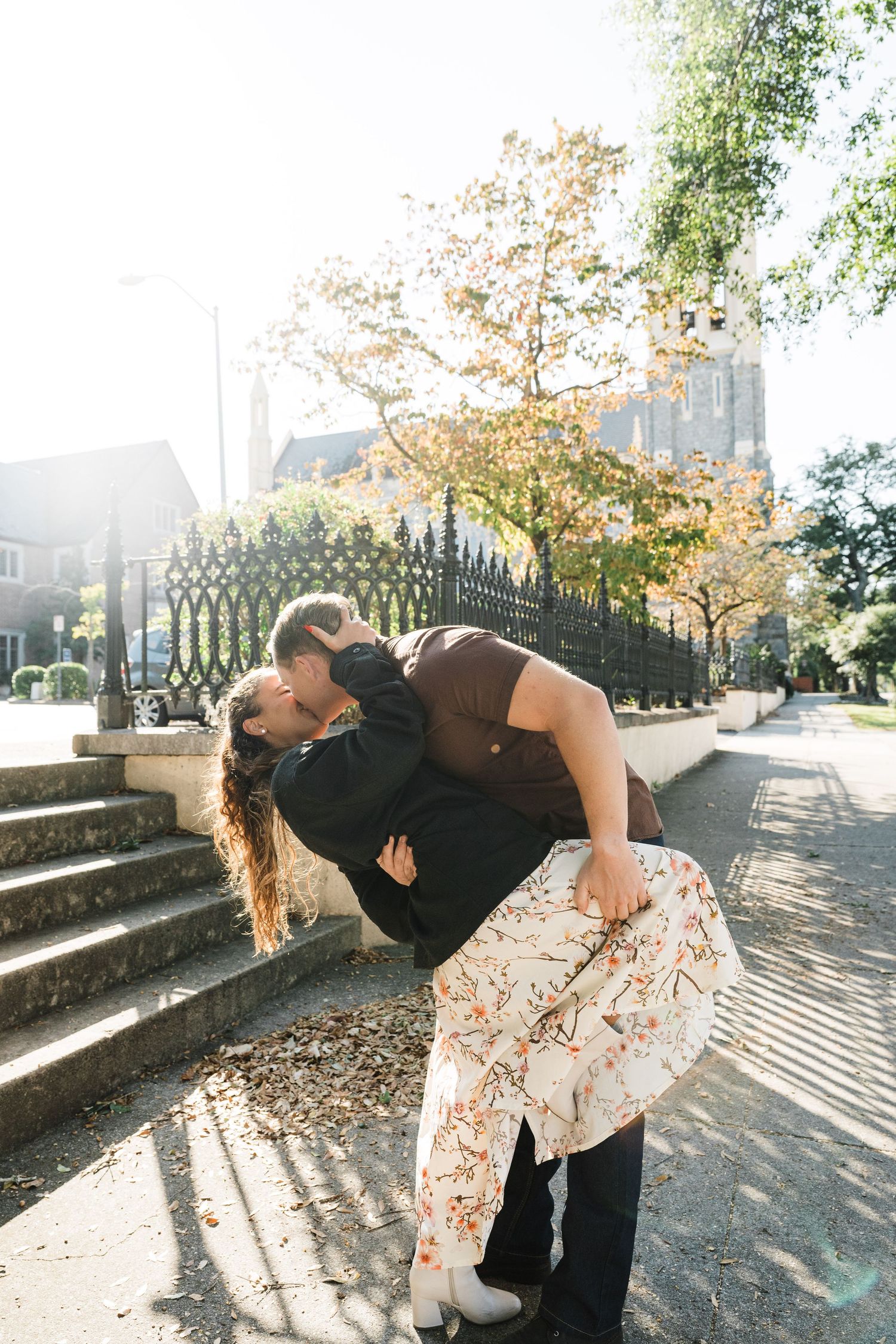 Romantic dip kiss between couple on sunlit steps with ornate iron fence and autumn leaves.
