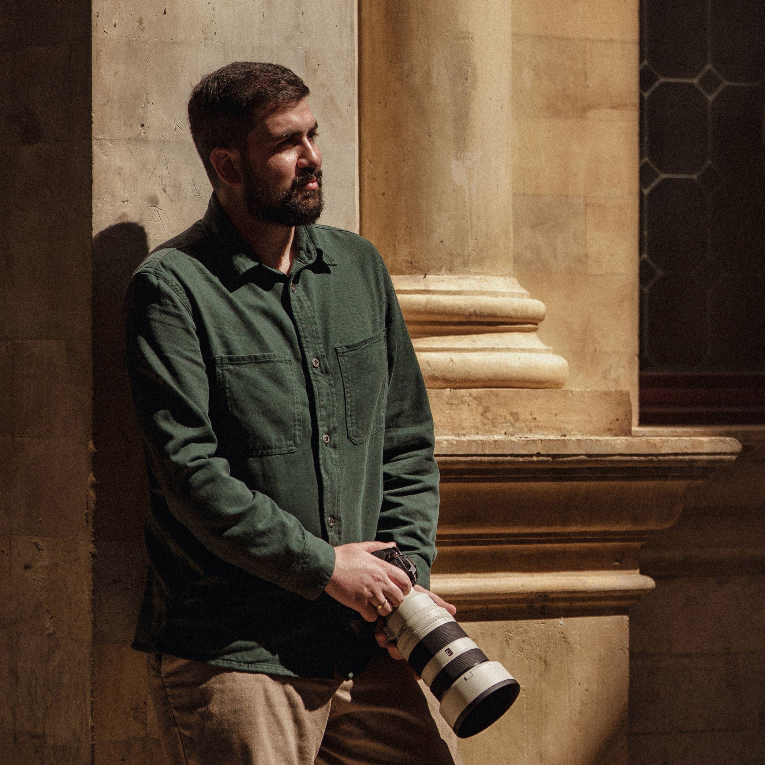 Photographer in grey sweater and leather straps holding camera in rustic wooden interior.