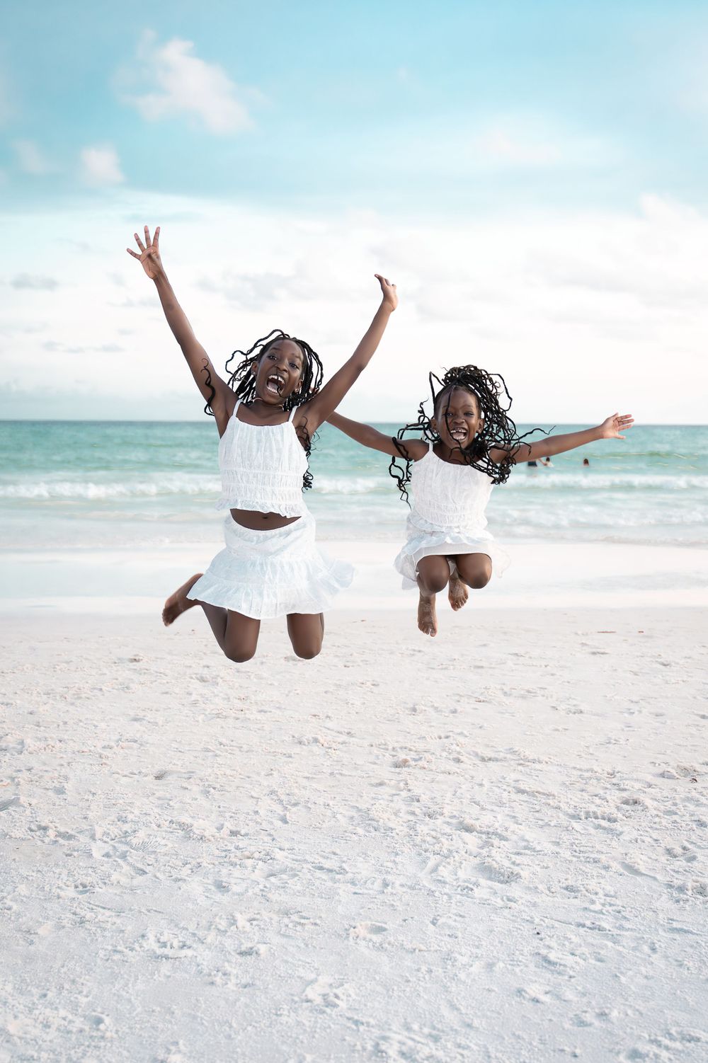 Two young girls in white dresses jumping joyfully on a sunny beach.