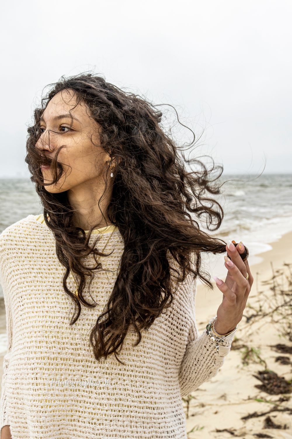 Woman with long dark curly hair laughing joyfully on a windy beach while wearing a cream knit sweater.