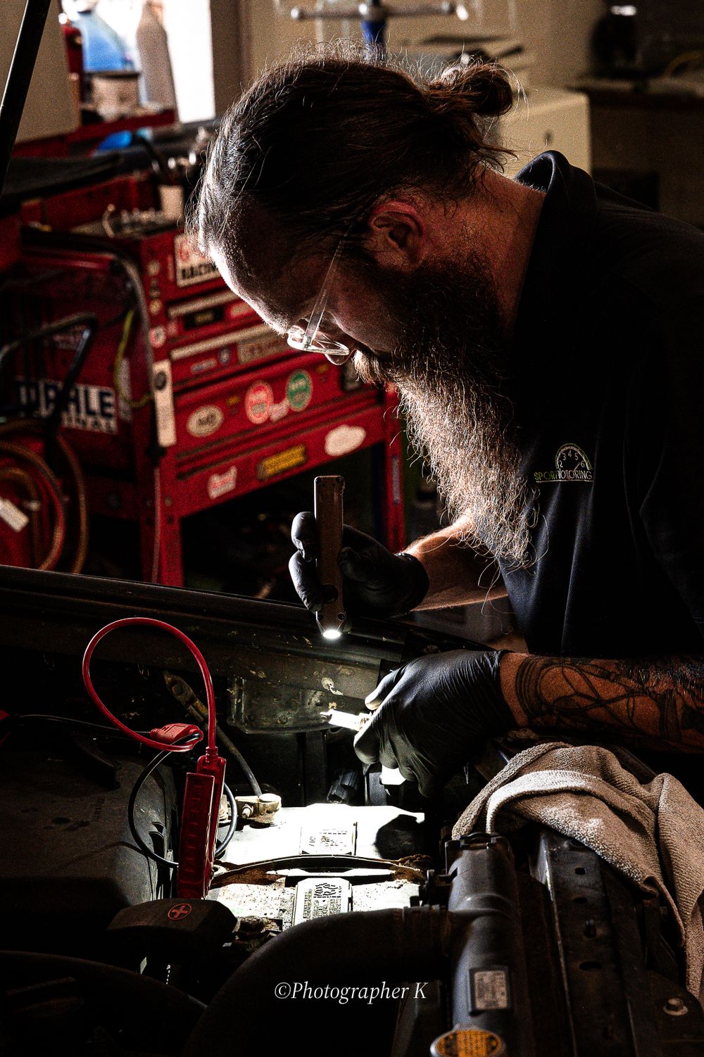 A bearded mechanic with glasses works intently in a dimly lit garage with vintage red toolboxes visible in background.