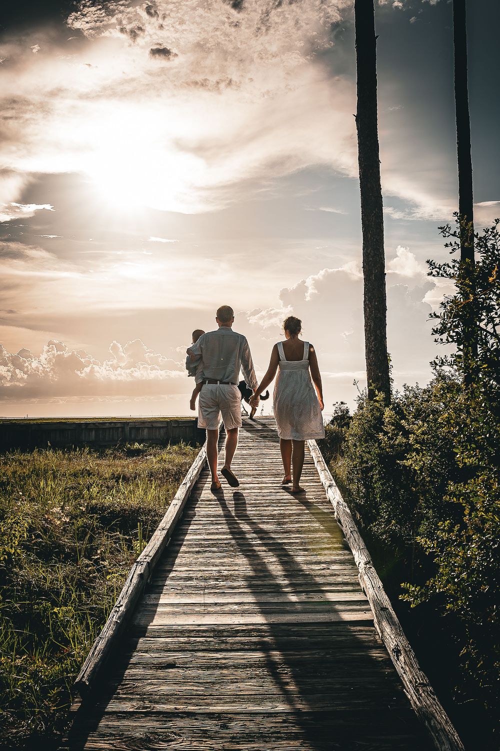 Two people walk along a wooden boardwalk leading to a cloudy horizon at sunset, creating dramatic silhouettes.