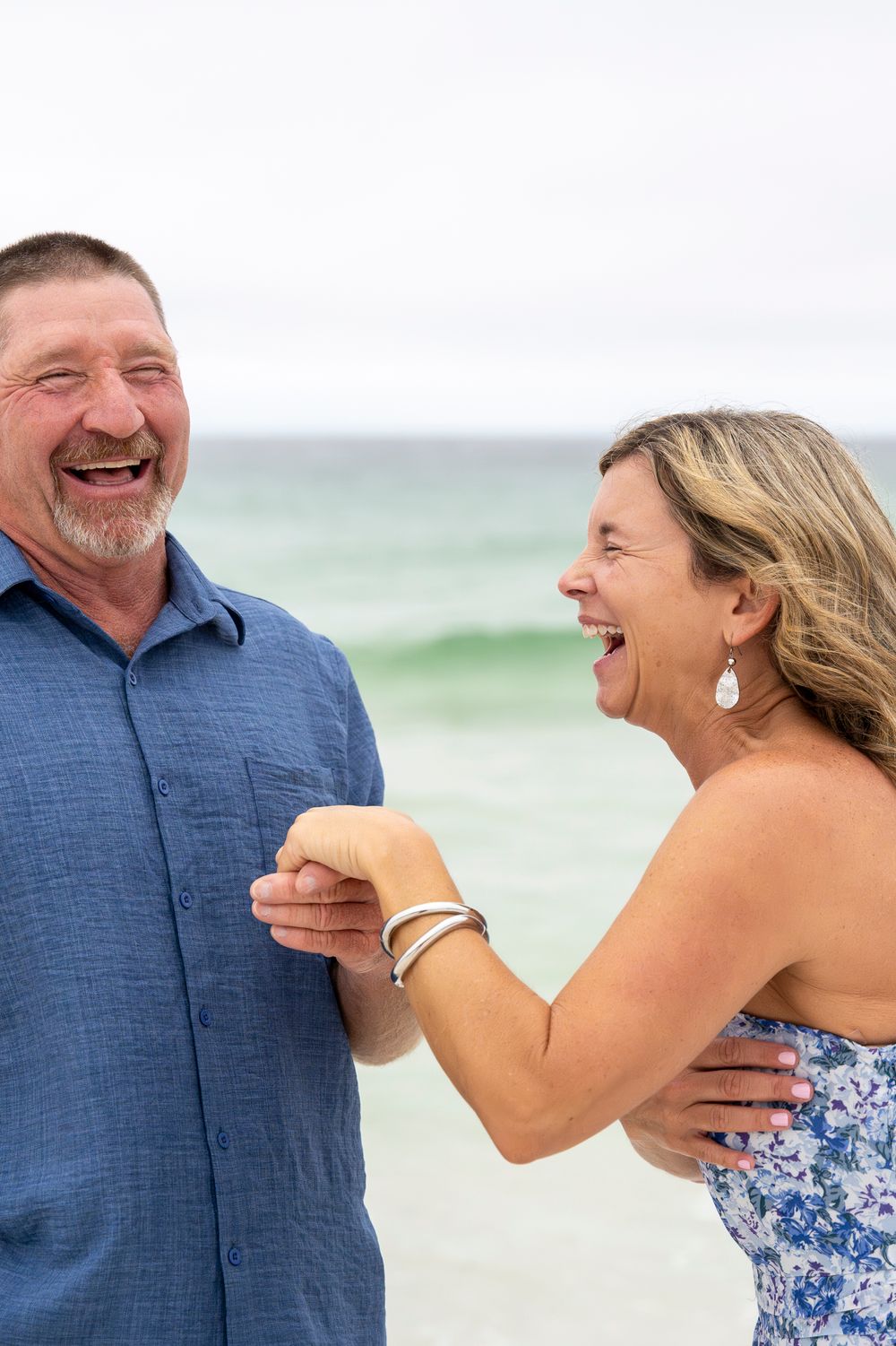 A couple shares joyful laughter while standing together on a beach with ocean waves in the background.