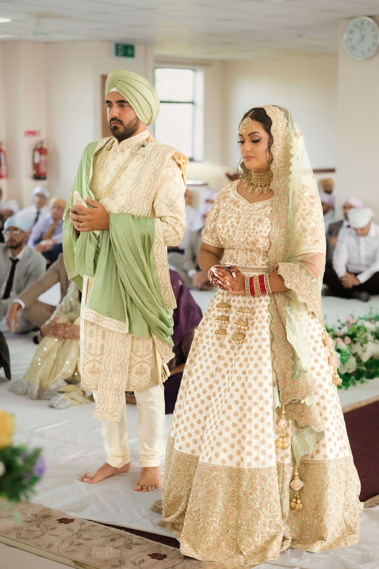 Sikh couple standing up in a Gurdwara receiving blessings from guru nanak whilst wearing light green asian attire
