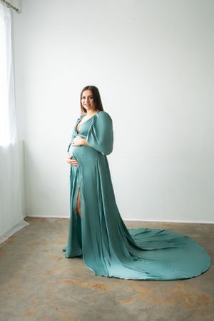 An elegant maternity portrait in a flowing teal dress with a dramatic train against a white background.