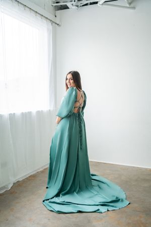 A flowing seafoam green maternity gown with an elegant open back design photographed in a minimalist studio setting.