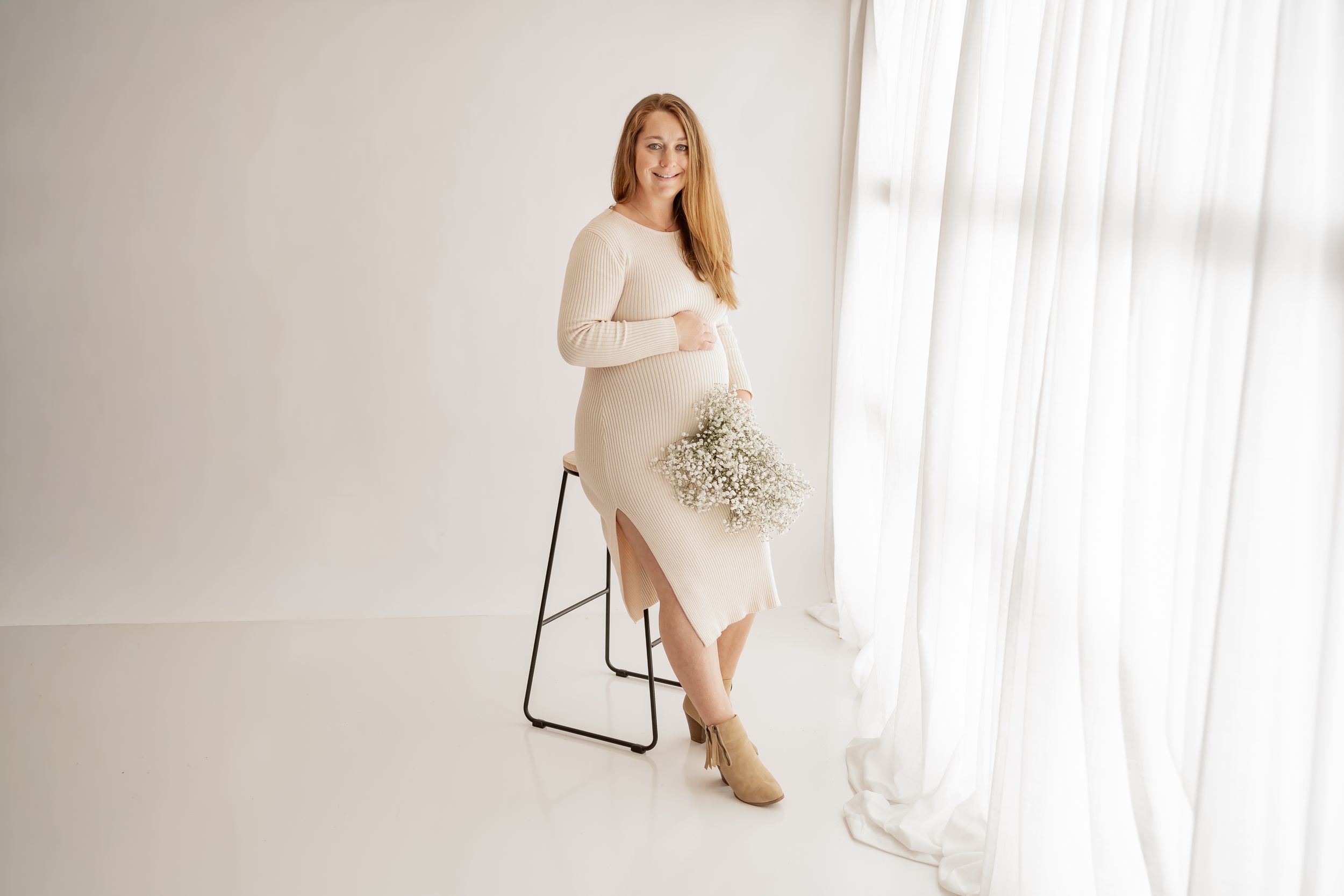 Elegant bride in white dress poses gracefully near bright window with soft natural light streaming in.