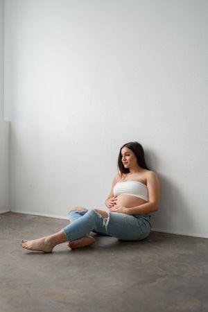 Expectant mother in casual attire sitting against a white wall on concrete floor.