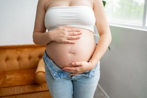 Close up of pregnant belly with hands resting on it, wearing white top and jeans.