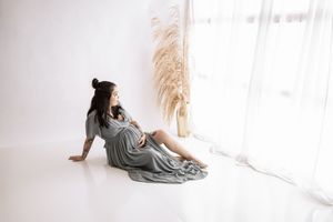 A person in a flowing sage green dress reclines on a white floor near pampas grass by a bright window.