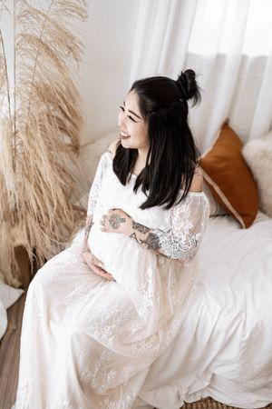 Woman in white lace dress lounges gracefully on white bedding with pampas grass decor in background.