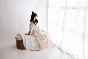 A person in a flowing white dress sits elegantly by a window in a minimalist white room with soft natural lighting.