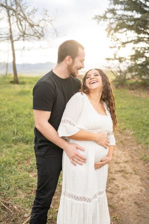Expectant couple embracing and laughing together on a sunlit nature path.