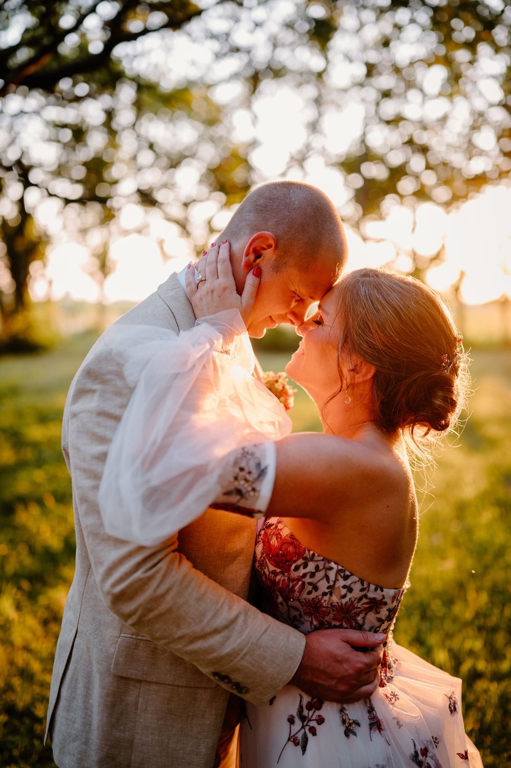 Romantic couple silhouetted against golden sunset in wedding attire share intimate moment in countryside meadow.