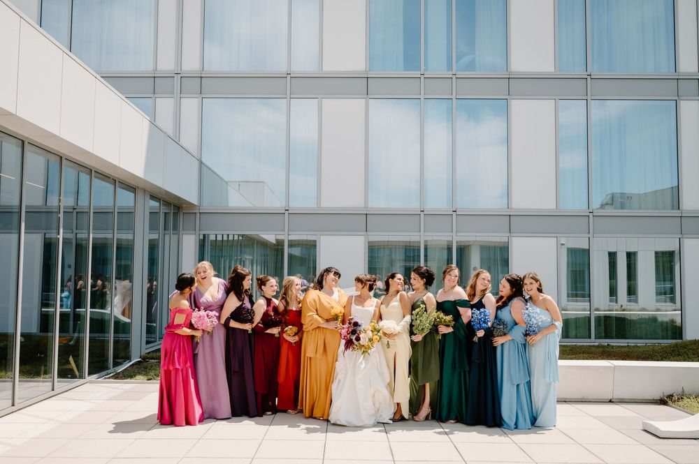 Wedding party in colorful dresses poses in front of modern glass building exterior.