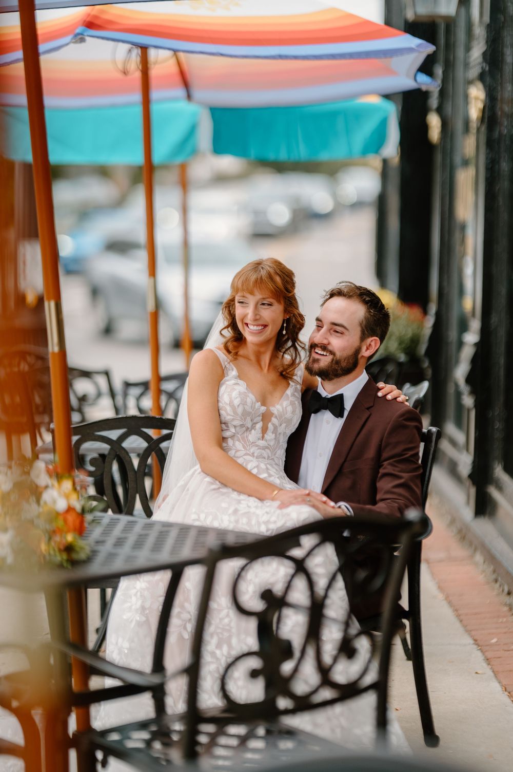 A couple in wedding attire sits together on a bench under a striped awning outside a cafe.