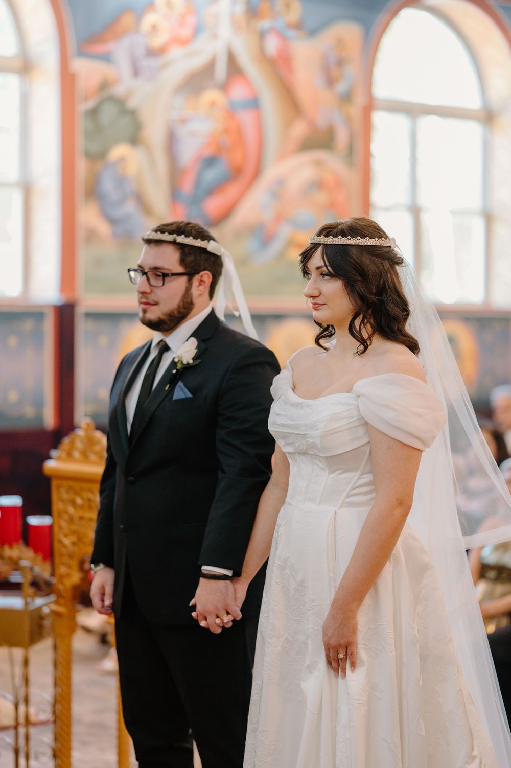A couple stands together during a wedding ceremony in a church with decorative wall murals in the background.