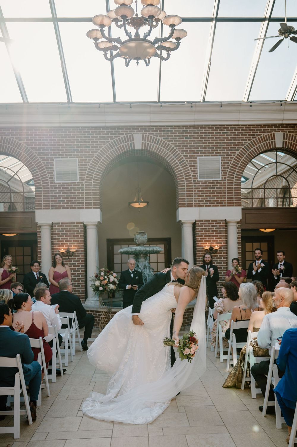 Wedding ceremony in a bright sunlit room with brick walls, arched windows, and white chairs arranged in rows.