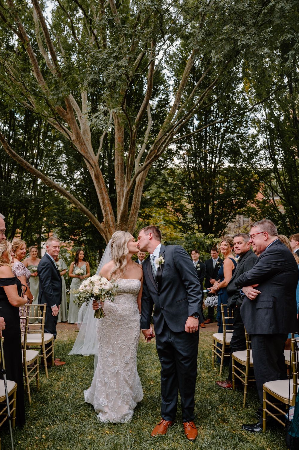 Guests at a woodland outdoor wedding ceremony rise from their chairs to greet the couple during a pivotal moment.