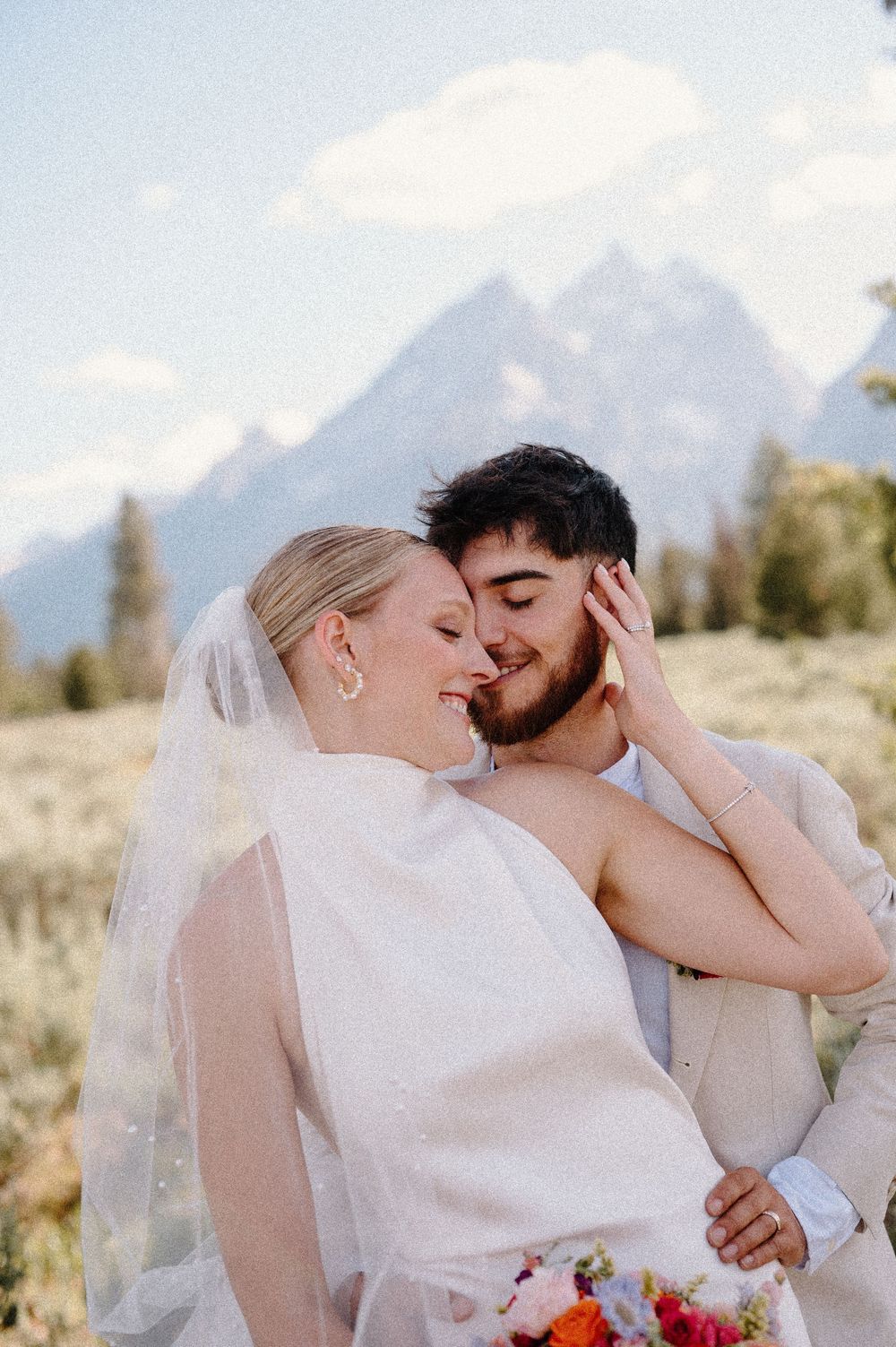 Two people share an intimate embrace outdoors against a scenic mountain landscape with blue skies.