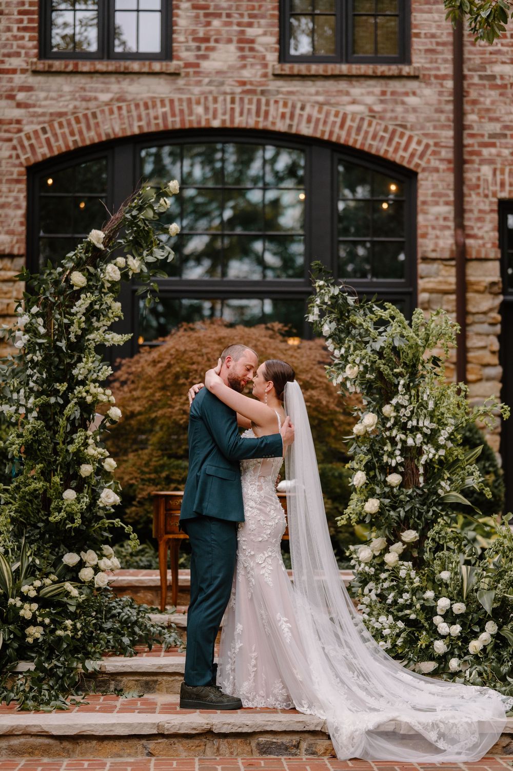 Romantic wedding couple sharing sweet moments against a rustic brick wall adorned with climbing white roses.