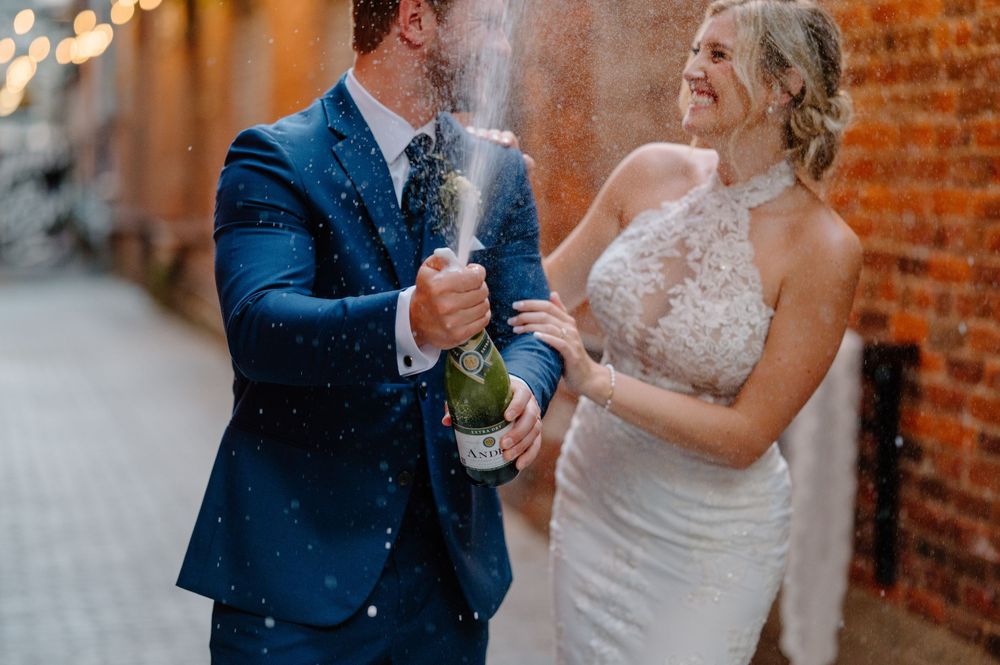 Couple in formal wedding attire dances together against a rustic brick wall backdrop in romantic lighting.