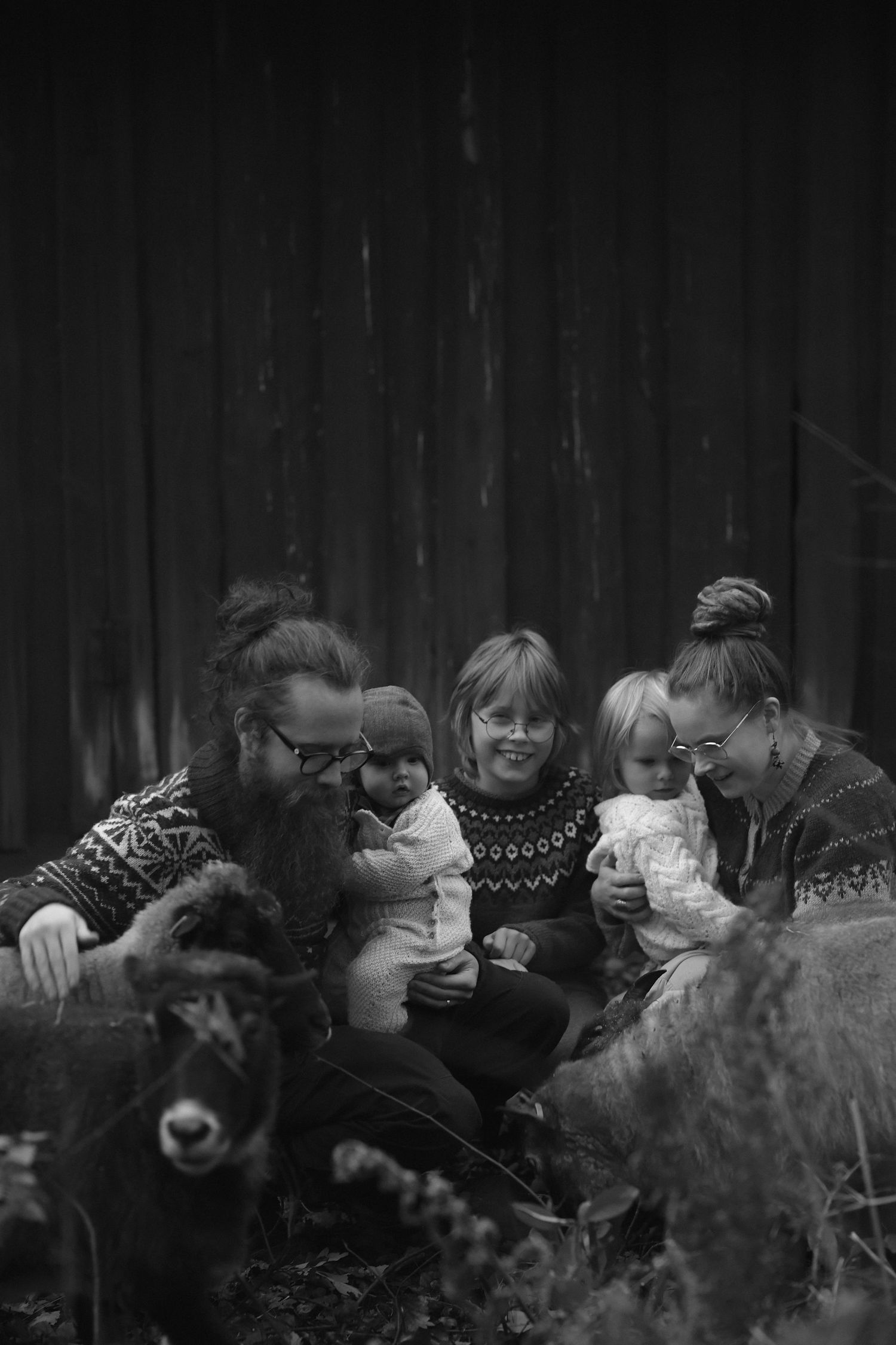 Black and white photo of a family in matching sweaters sitting together outdoors with their sheep.