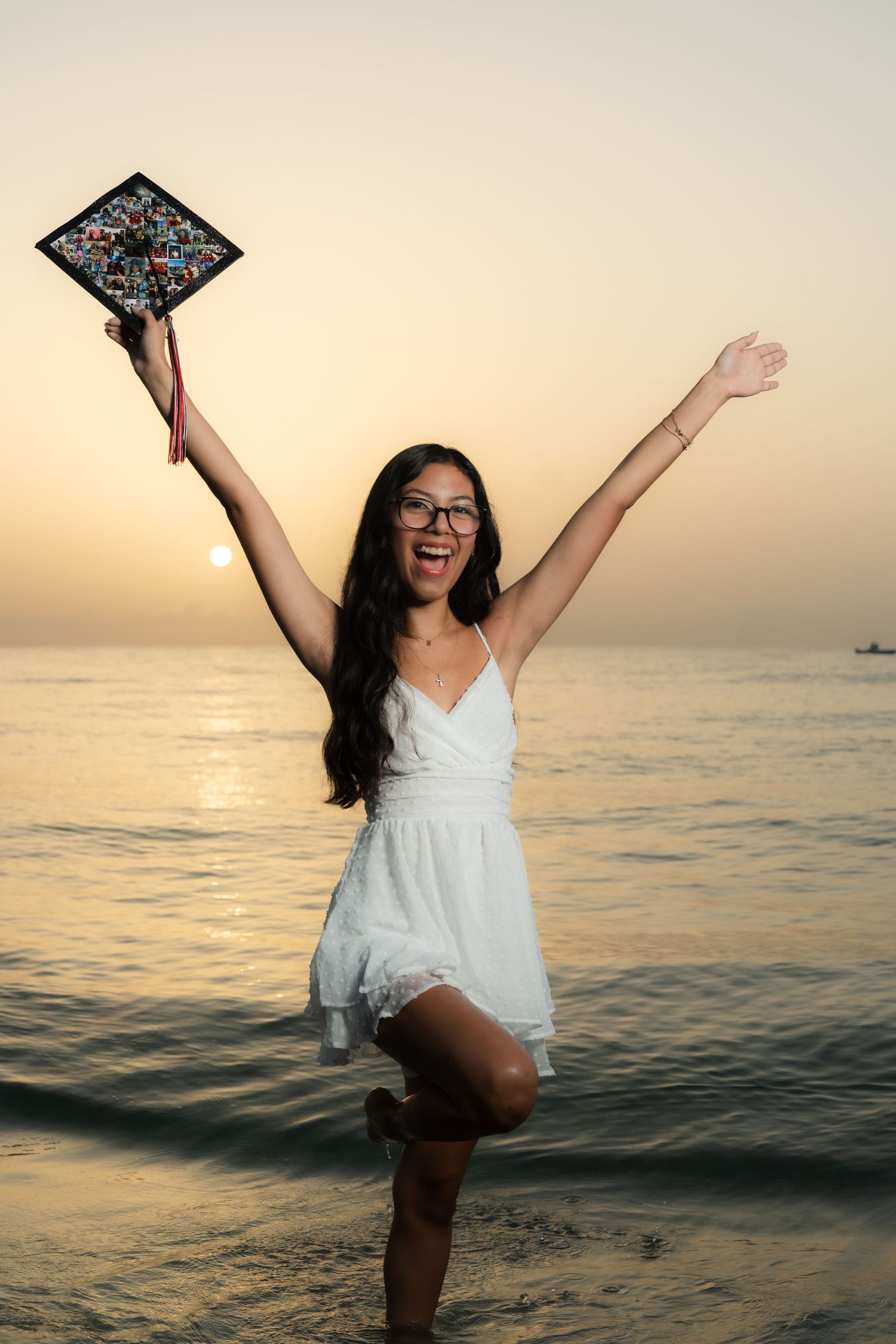 Graduate celebrates in white dress at sunset on beach holding decorated cap with outstretched arms.